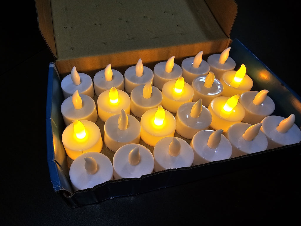 A set of 16 white LED tea light candles with glowing yellow flames, arranged in a grid inside an open cardboard box on a dark background.