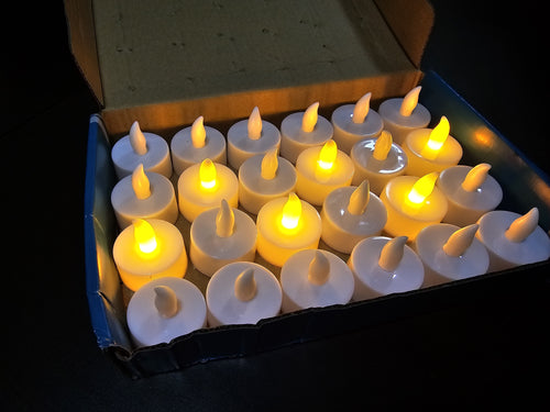 A set of 16 white LED tea light candles with glowing yellow flames, arranged in a grid inside an open cardboard box on a dark background.